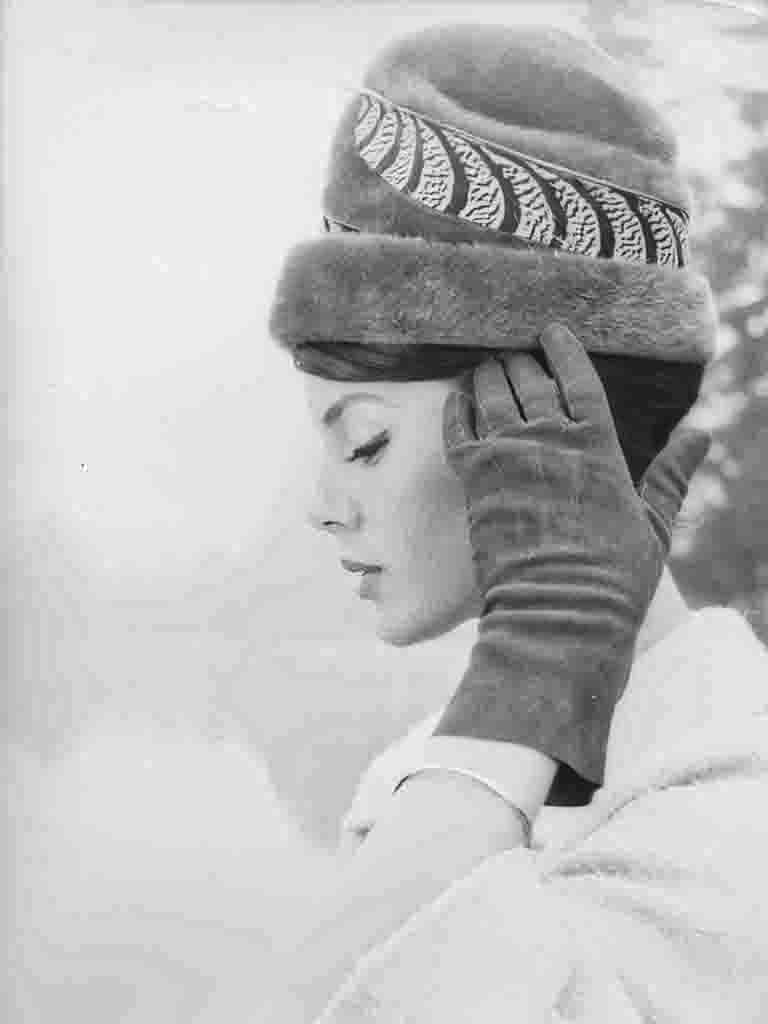 Eine kunstvolle Profilaufnahme, die ganz im Zeichen der Accessoires steht. Der hohe Hut mit Pelzbesatz und grafischem Muster bildet zusammen mit den dunklen Wildlederhandschuhen ein harmonisches Ensemble. Die helle, fast traumartige Belichtung lenkt die gesamte Aufmerksamkeit auf die Texturen der Mode.